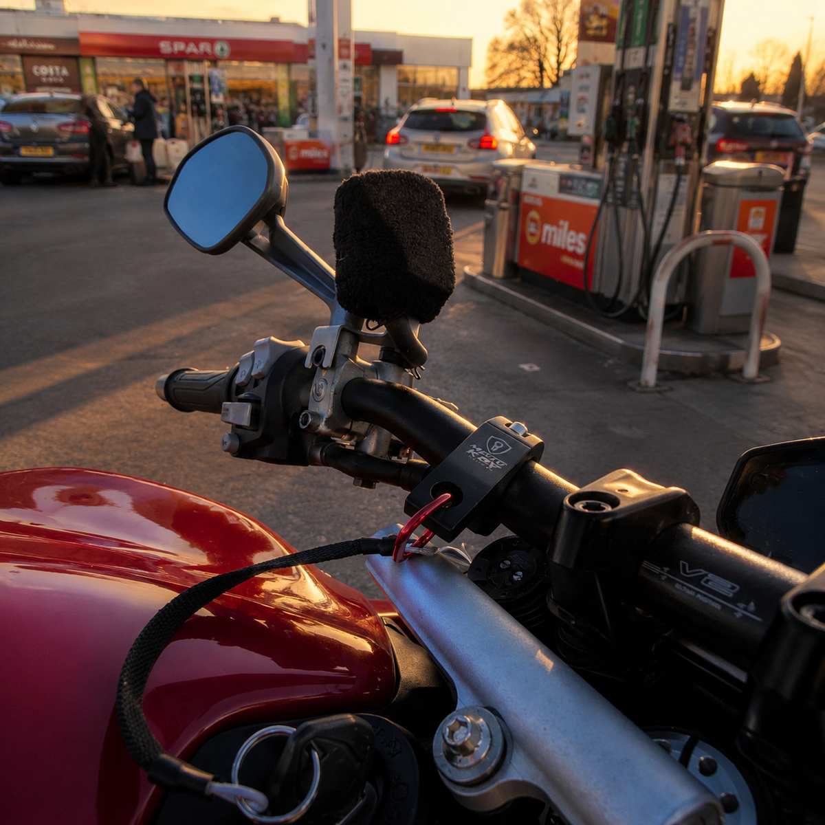 Close-up of a motorcycle handlebar with Motokey Guard setup at a gas station during golden hour, with a black foam windshield protector mounted on the mirror area. Visible brands include X-Cell on the handlebar clamp and SPAR6 on the gas station sign in the background.